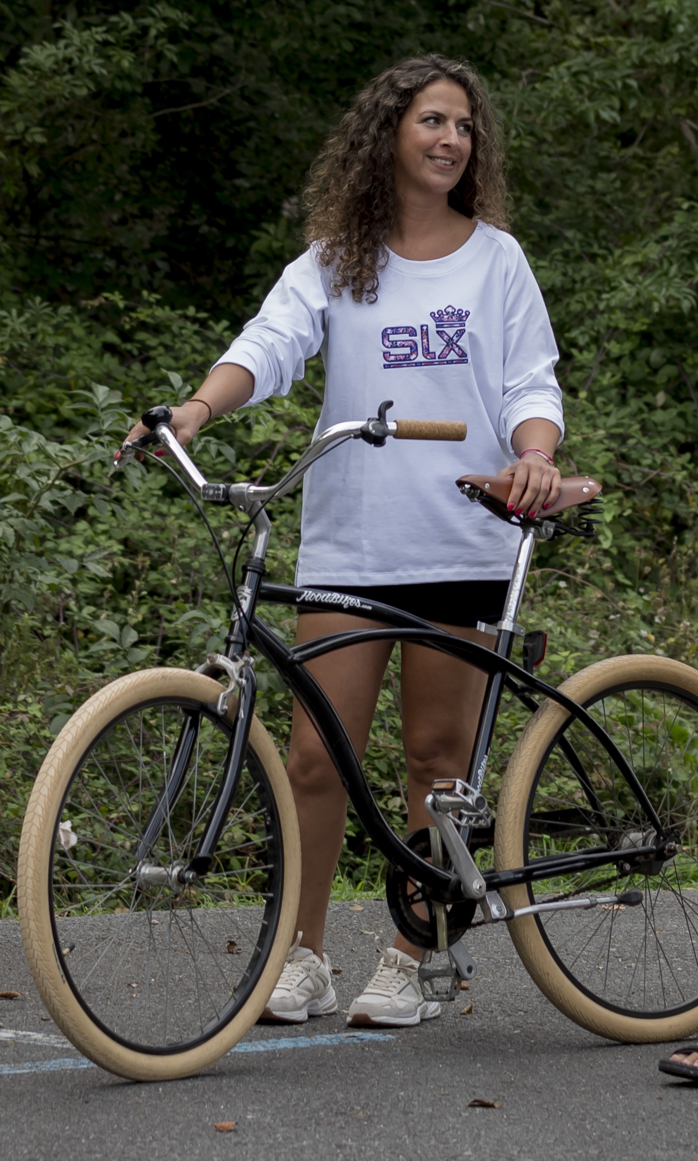 CHICA CON UNA BICICLETA VINTAGE , UN PANTALON CORTO Y UNA SUDADERA BLANCA CON LA MARCA BORDADA EN EL PECHO CON UN ESTAMPADO FLORAL.