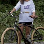 CHICA CON UNA BICICLETA VINTAGE , UN PANTALON CORTO Y UNA SUDADERA BLANCA CON LA MARCA BORDADA EN EL PECHO CON UN ESTAMPADO FLORAL.