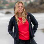 mujer con chaqueta marino y capucha puesta , con una camiseta roja en la orilla del mar