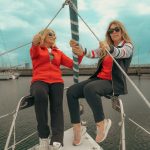 dos mujeres en la proa de un barco vestidas con pantalones marinos y chaquetas roja y marino.