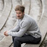 HOMBRE SENTADO EN UN ESCALON CON SUADERA GRIS CLARA Y PANTALON GRIS OSCURO