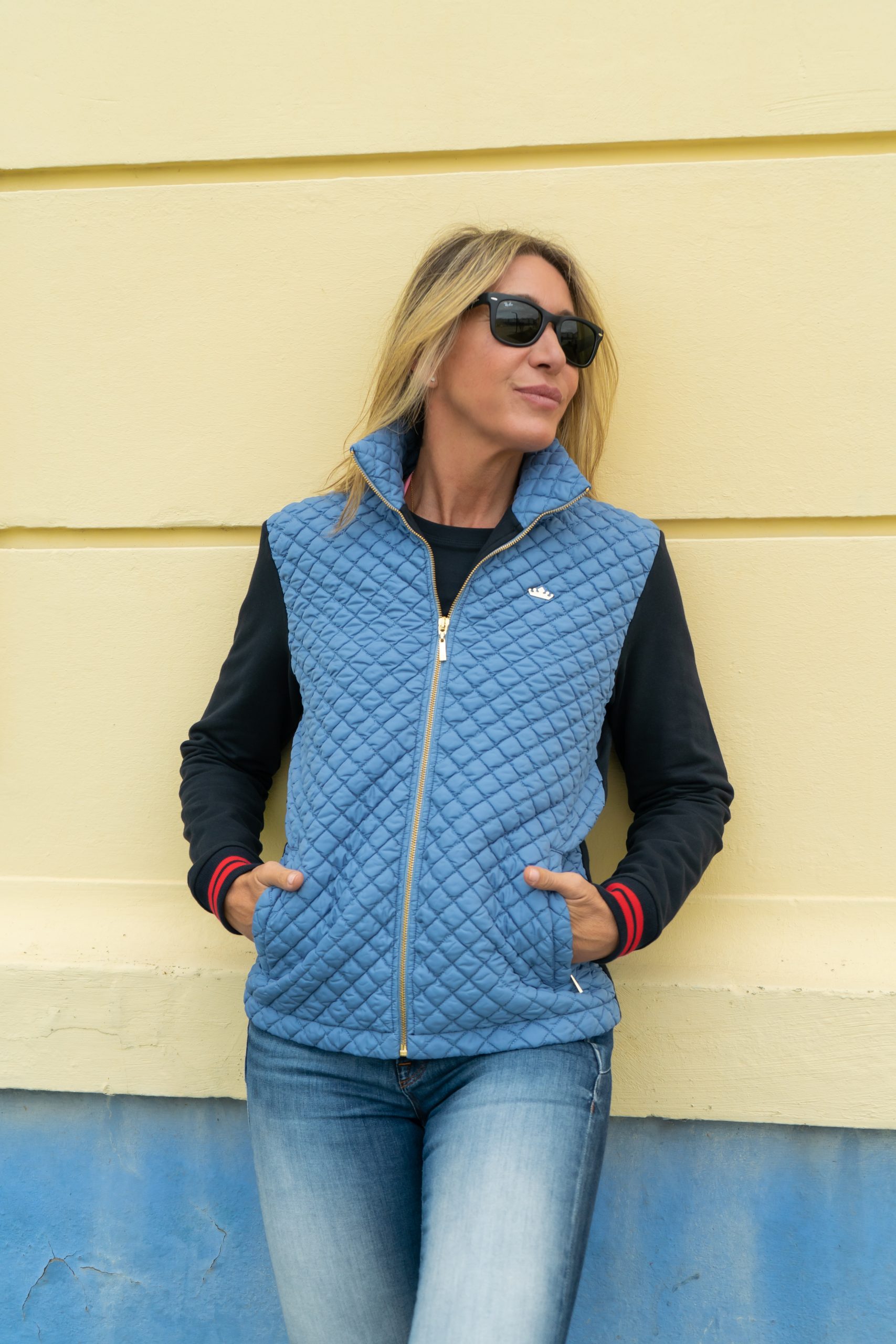 una mujer aployada en una pared amarilla con una chaqueta acolchada celeste con las mangas marinas.