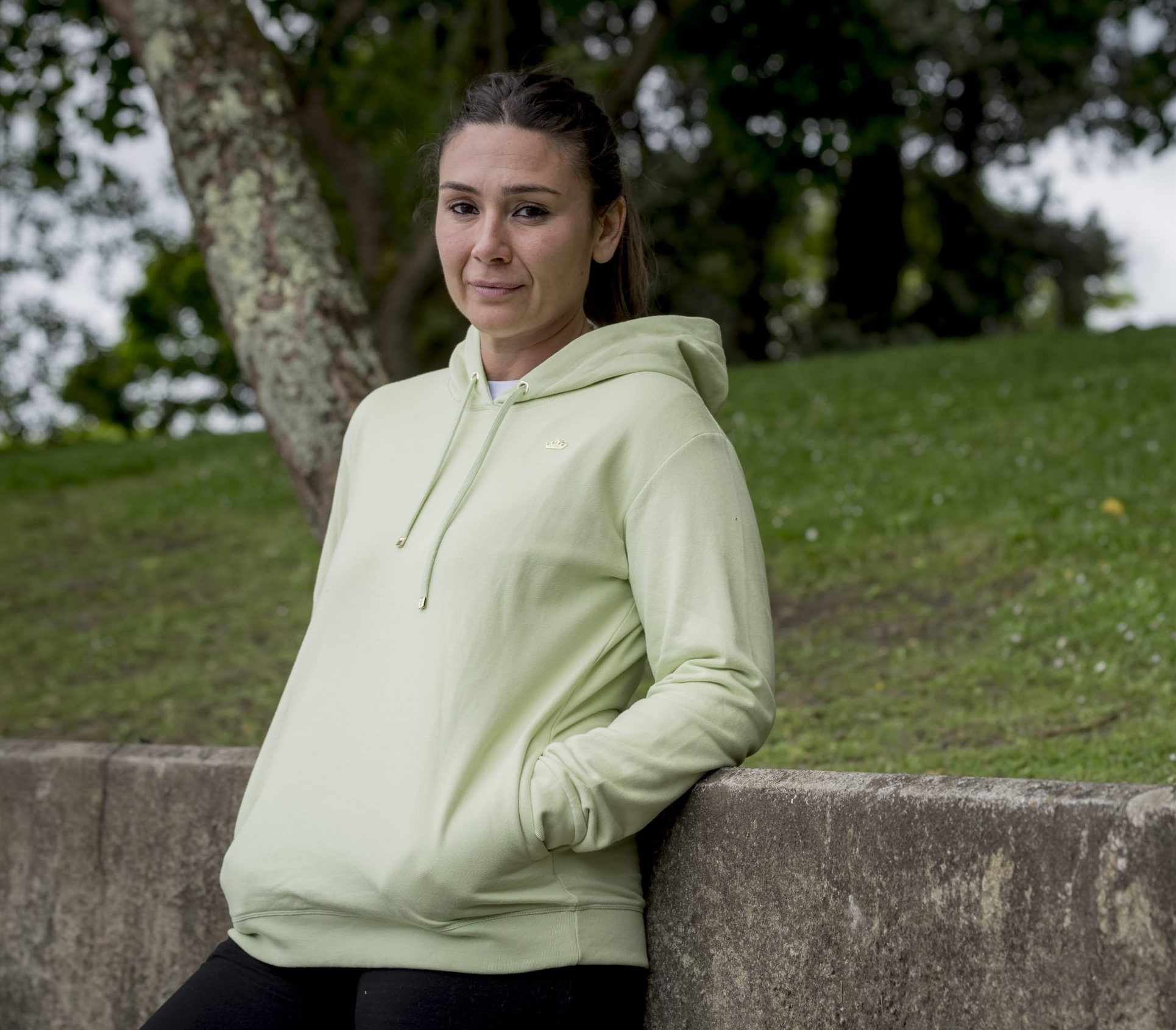 chica con una sudadera en color matcha apoyada a un muro de piedra en un parque.
