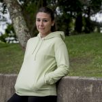 chica con una sudadera en color matcha apoyada a un muro de piedra en un parque.