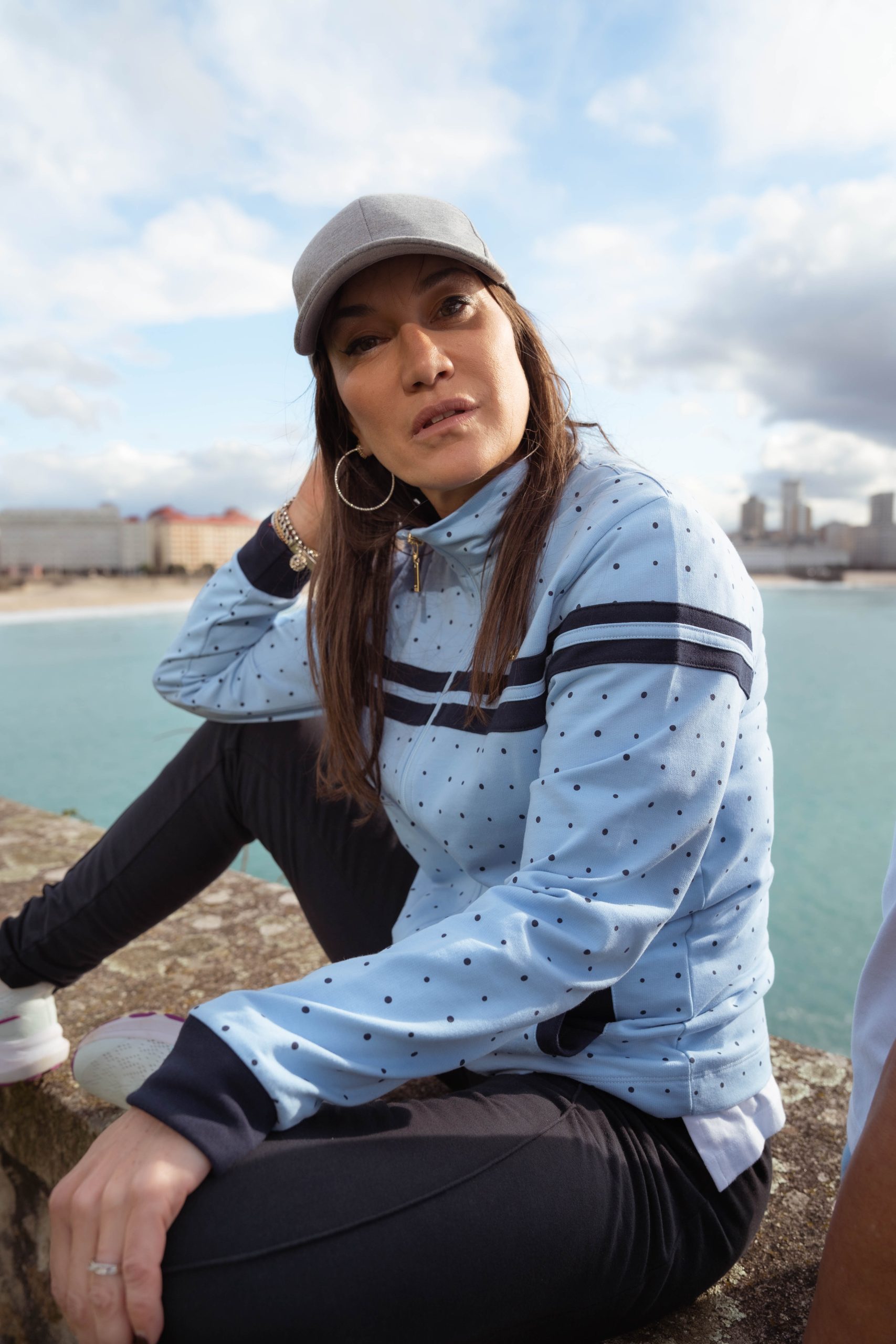 CHICA SENTADA EN UN MURO CON EL MAR DE FONDO Y UNA CHAQUETE CELESTE MUDEJAR CON DETALLES EN COLOR MARINO TNTO LUNARES , COMO PUÑOS.