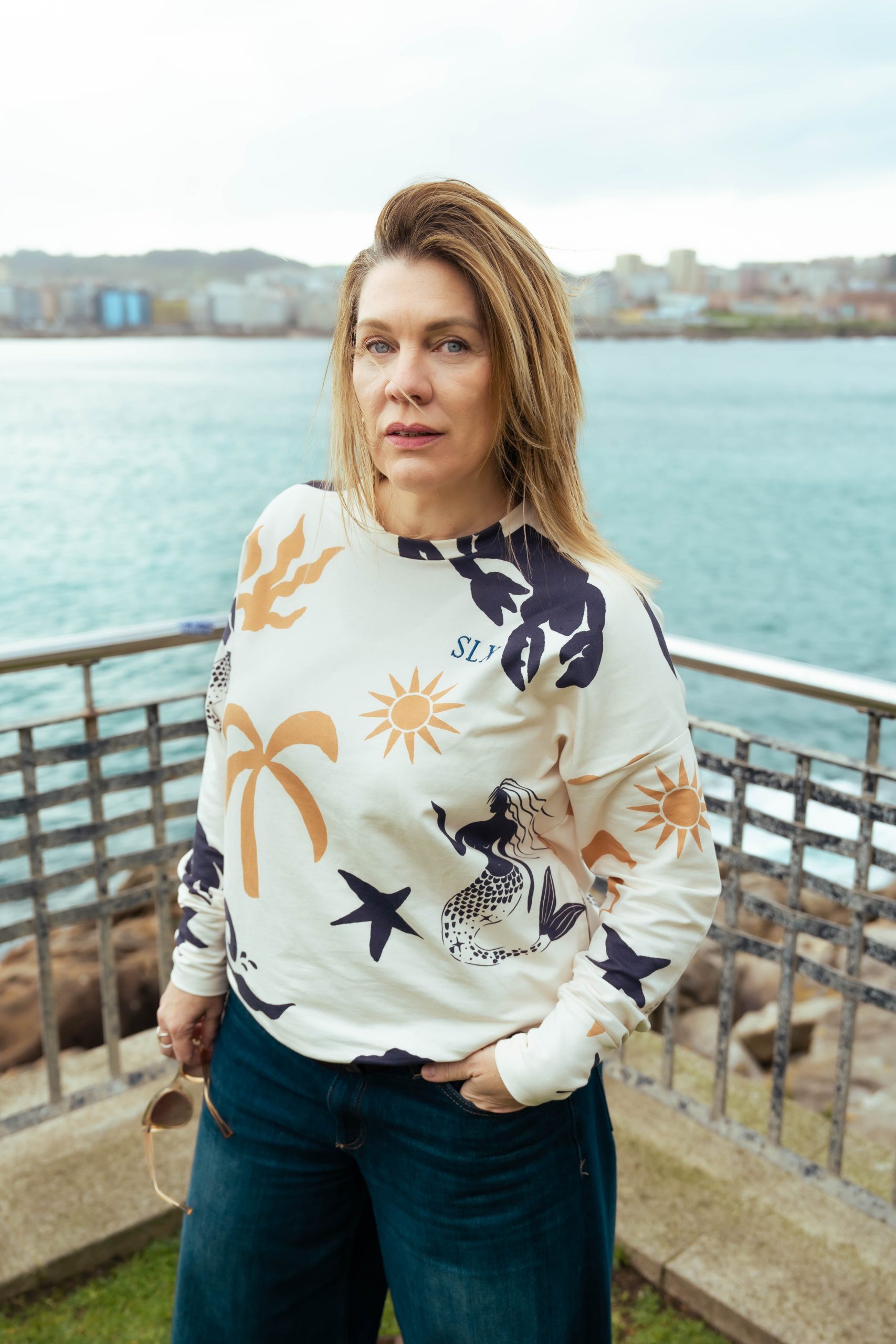 chica en una barandilla con el mar de fondo y nuestra sudadera sirena con un estampado super veraniego