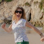 MUJER CORRIENDO EN LA ORILLA DE LA PLAYA DE MANERA DIVERTIDA CON UN PAMTALON VERDE Y UNA CAMISETA BLANCA CON LUNARES VERDE Y LAVANDA.