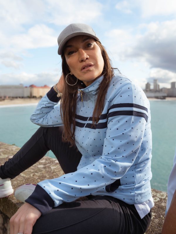 CHICA SENTADA EN UN MURO CON EL MAR DE FONDO Y UNA CHAQUETE CELESTE MUDEJAR CON DETALLES EN COLOR MARINO TNTO LUNARES , COMO PUÑOS.