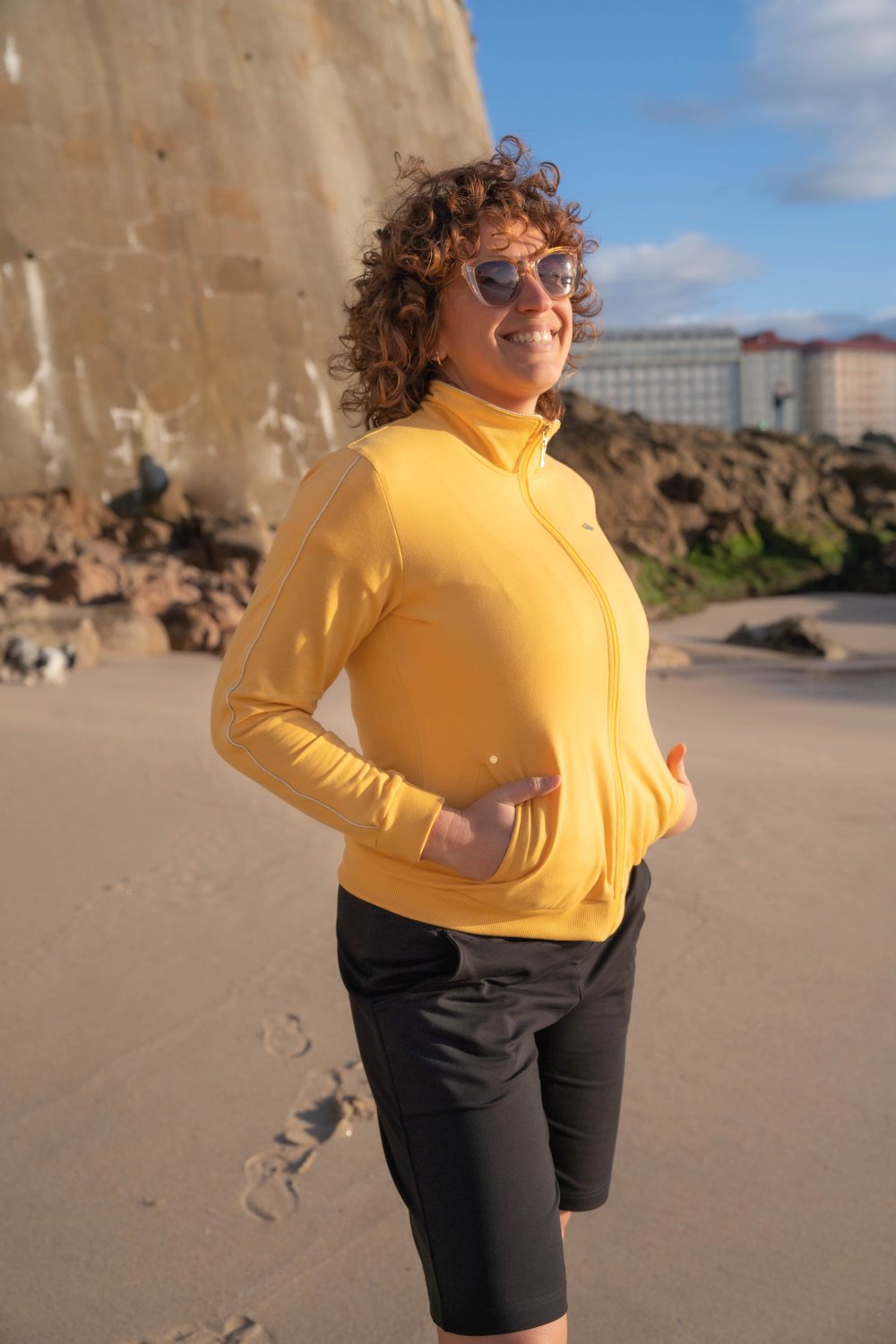 una mujer mirando al mar con gafas de sol, manos en los bolsillos y lleva puesta una chaqueta amarilla