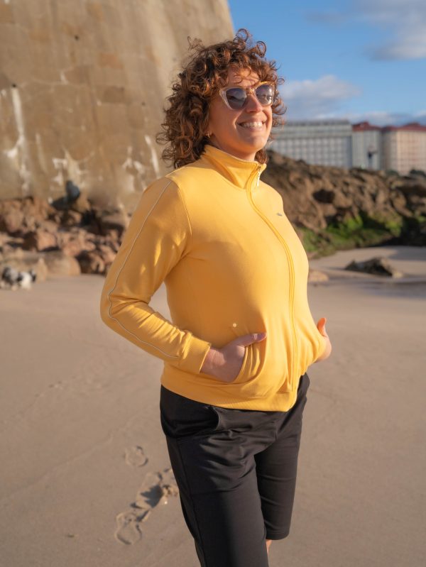 una mujer mirando al mar con gafas de sol, manos en los bolsillos y lleva puesta una chaqueta amarilla