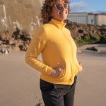 una mujer mirando al mar con gafas de sol, manos en los bolsillos y lleva puesta una chaqueta amarilla