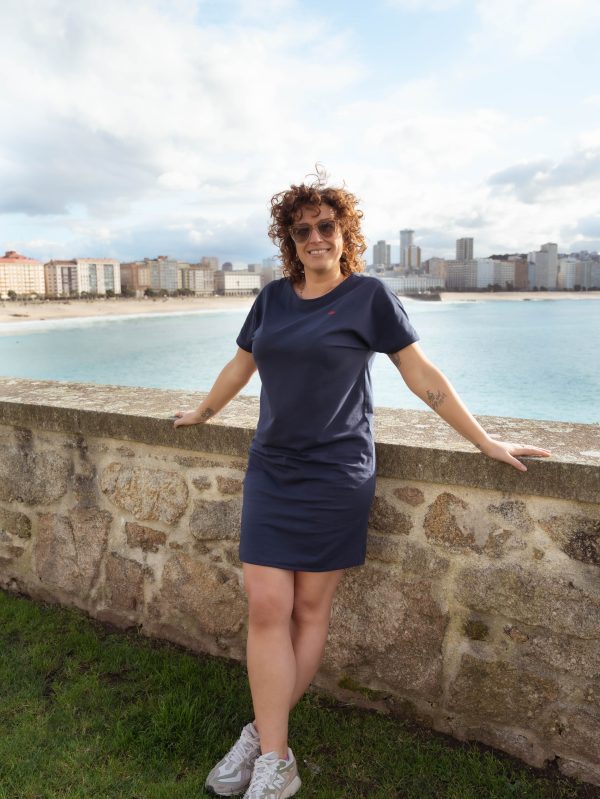 CHICA APOYADA EN UN MURO DE PIEDRA CON EL MAR DE FONDO Y UN VESTIDO EN COLOR MARINO CON MANGAS RANGLAN .